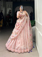 Model showcasing a premium baby pink metallic tissue silk Chaniya Choli in a saree-drape style, featuring a wide solid border skirt, multi-color pastel floral vine embroidery on the dupatta, and hanging purple tassels inside a boutique showroom.