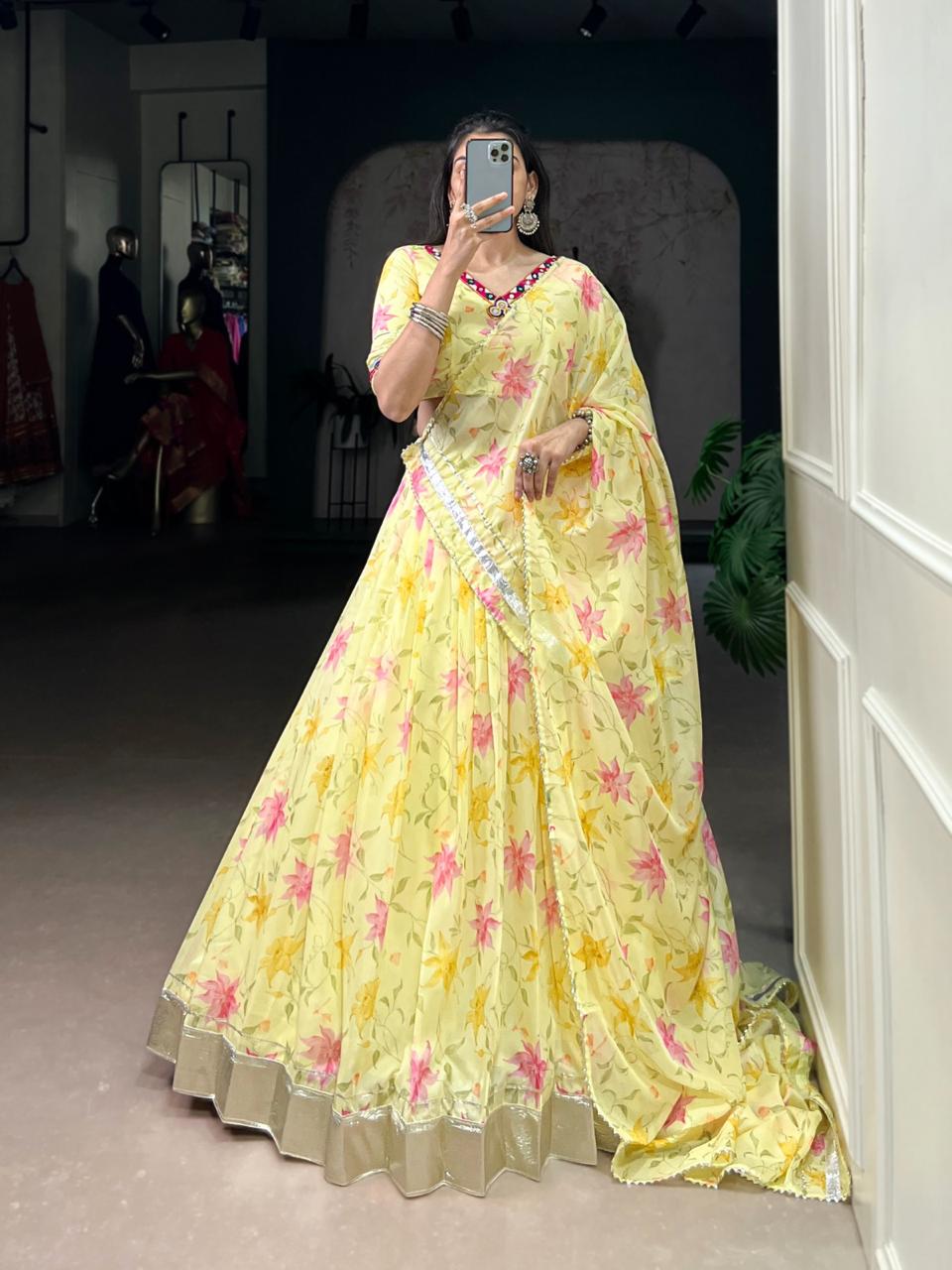 Model showcasing a premium canary yellow georgette Chaniya Choli with a vibrant pink watercolor floral print, a wide silver gota tissue border, and a matching embroidered V-neck blouse inside a boutique. Main image