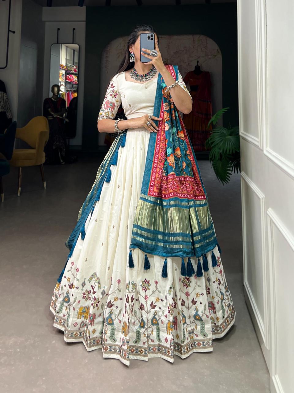 Model showcasing a premium off-white silk Chaniya Choli featuring a traditional multi-color Kalamkari peacock print border and a heavy teal Patola print dupatta with gold zari work inside a boutique. Main image