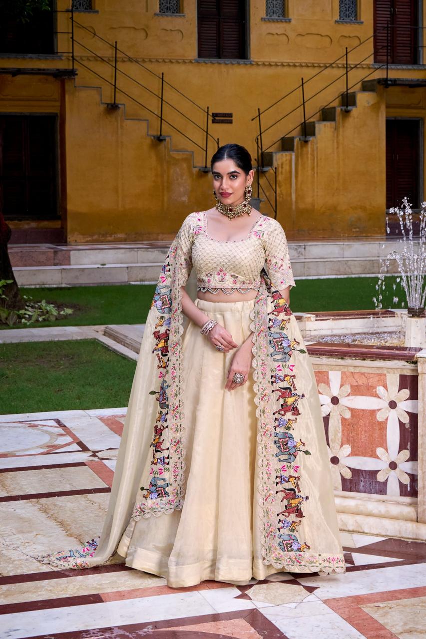 Model showcasing a premium cream organza silk Chaniya Choli featuring a clean fluid skirt, a pearl-beaded floral blouse, and a majestic sheer dupatta bordered with a colorful folk-art embroidery depicting a royal elephant procession inside a courtyard setting. Main image