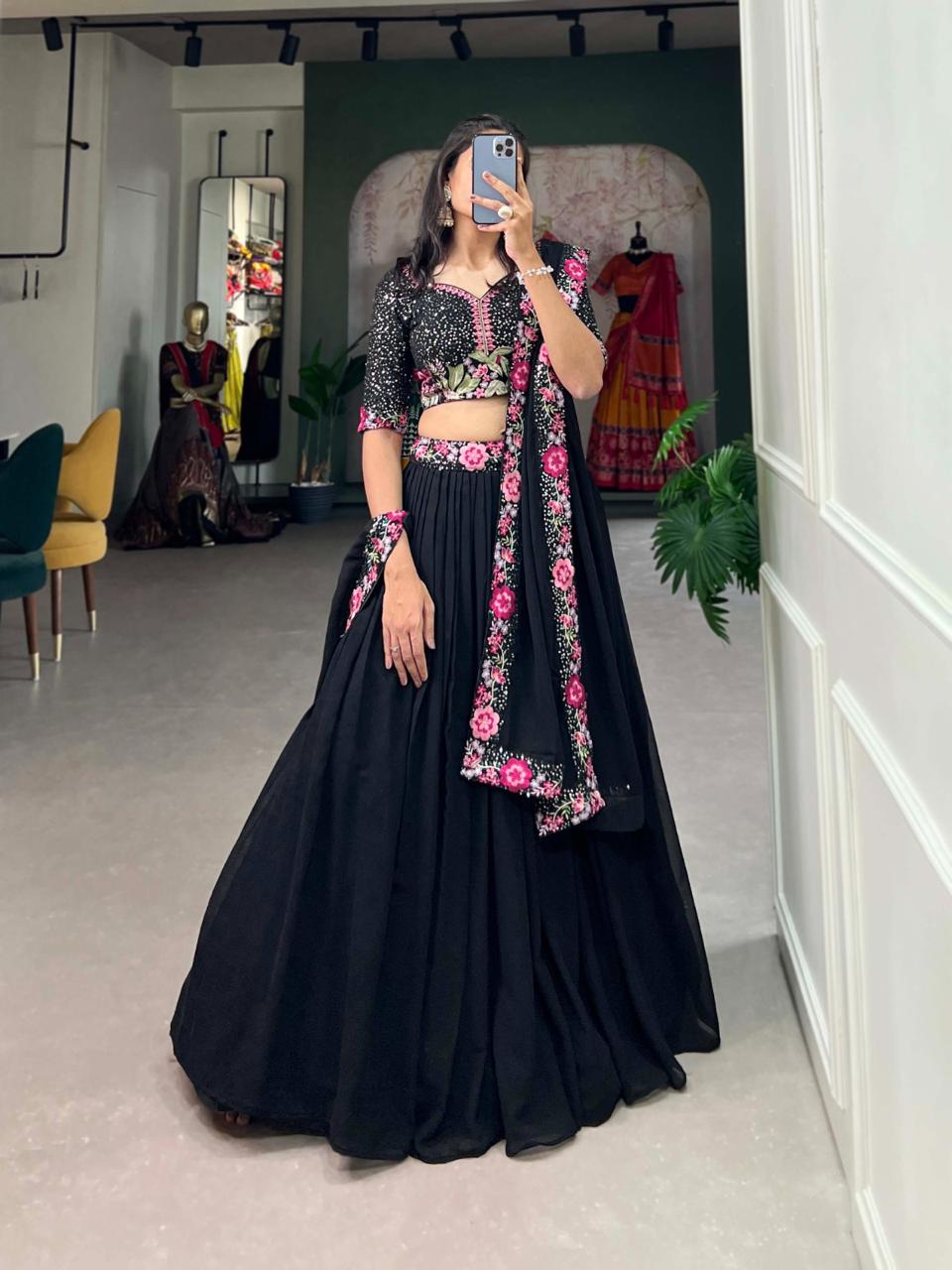 Model showcasing a premium midnight black georgette Chaniya Choli featuring a clean fluid skirt, a heavily embroidered pink floral and mirror work blouse, and a matching scalloped border dupatta inside a boutique showroom.