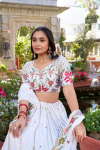 Model showcasing a premium off-white georgette Chaniya Choli featuring vertical gold zari lines on the skirt, heavily detailed multi-color floral resham embroidery on the blouse, and a matching scalloped floral border dupatta inside an outdoor garden.