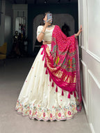 Model showcasing a premium off-white silk Chaniya Choli featuring multi-color floral hem embroidery and waist tassels, paired with a heavy vibrant pink silk dupatta with gold zari panels inside a boutique.