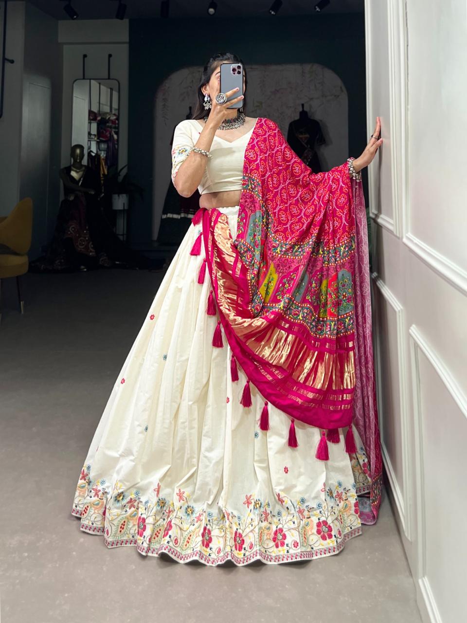 Model showcasing a premium off-white silk Chaniya Choli featuring multi-color floral hem embroidery and waist tassels, paired with a heavy vibrant pink silk dupatta with gold zari panels inside a boutique.