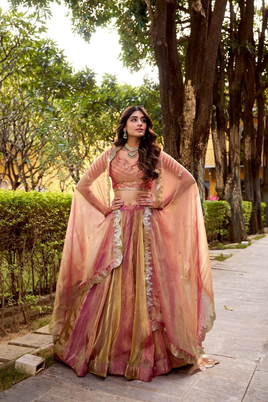 Model showcasing a premium peach pink and gold metallic tissue silk Chaniya Choli featuring a dual-tone shimmer, matching sleeveless choli blouse, and a heavy scalloped border dupatta worn in a cape style inside an outdoor garden. Main image