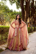 Model showcasing a premium peach pink and gold metallic tissue silk Chaniya Choli featuring a dual-tone shimmer, matching sleeveless choli blouse, and a heavy scalloped border dupatta worn in a cape style inside an outdoor garden.