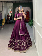 Model showcasing a premium deep wine silk Chaniya Choli featuring heavy gold zari and dori floral vine border embroidery, an intricately detailed matching mid-sleeve blouse, and a scalloped border dupatta inside a boutique showroom.
