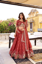 Model showcasing a premium brick red silk Chaniya Choli featuring an intricate traditional gold zari and resham palace-arch jharokha border embroidery, a matching heavily detailed blouse, and an embroidered dupatta inside a traditional heritage palace location.