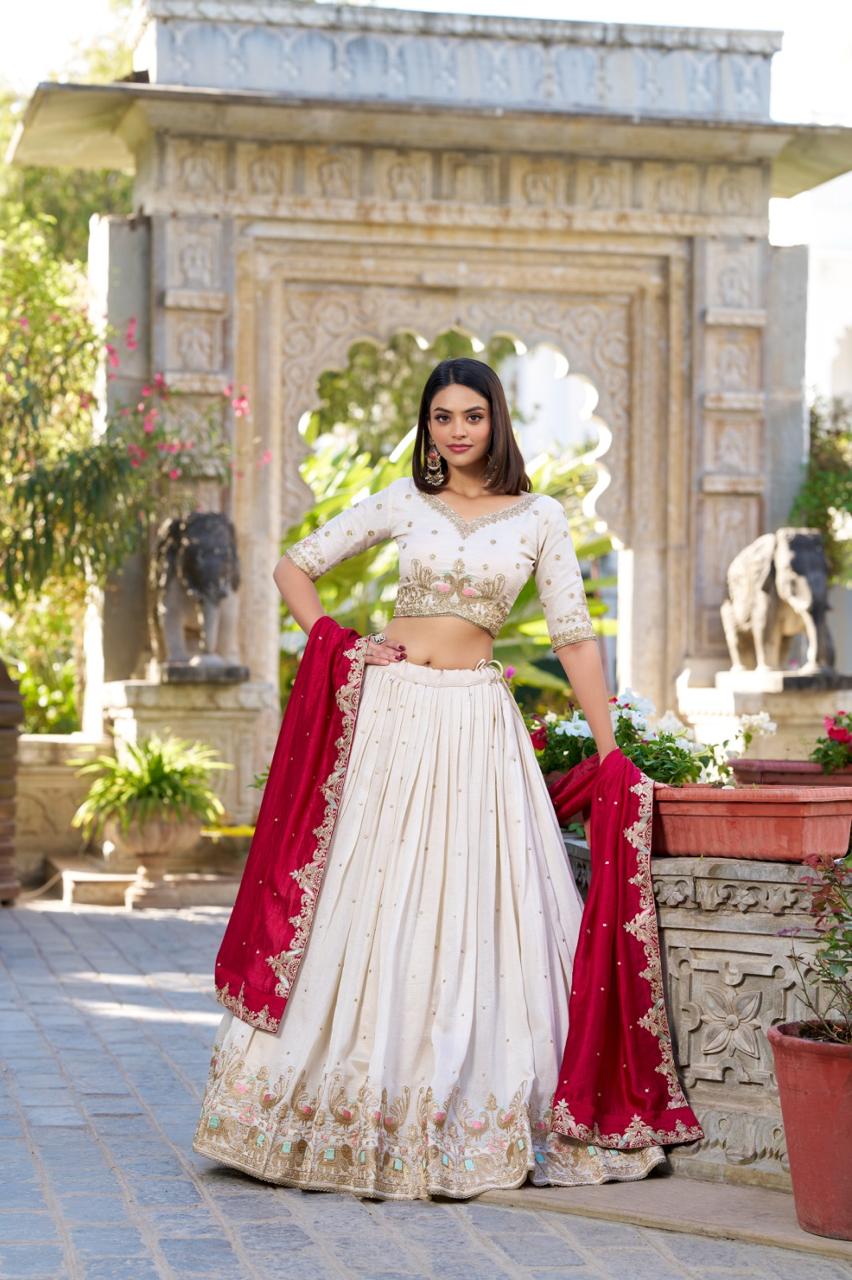 Model showcasing a premium ivory silk Chaniya Choli featuring a wide gold zari and resham peacock embroidered border, paired with a heavily detailed matching mid-sleeve blouse and a contrasting crimson red dupatta inside an outdoor traditional heritage setting.