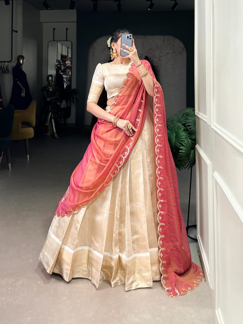 Model showcasing a premium ivory tissue silk Chaniya Choli in a saree-drape style, featuring a wide gold zari border skirt, an elegant elbow-sleeve blouse, and a coral pink organza dupatta with a scalloped gold zari border inside a boutique showroom.