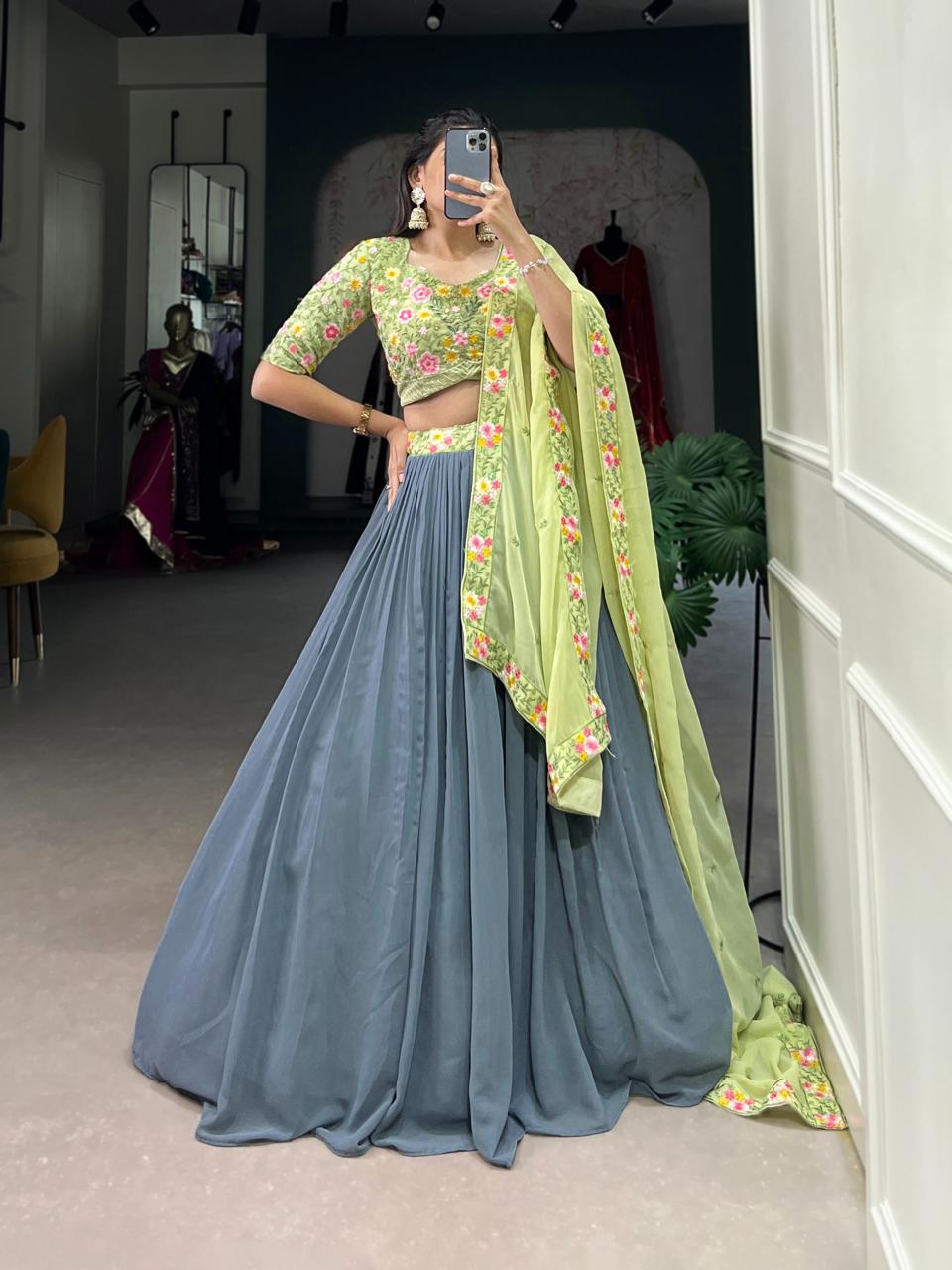 Model showcasing a premium slate grey georgette Chaniya Choli featuring a clean fluid skirt, paired with a heavily embroidered pistachio green floral blouse and matching embroidered border dupatta inside a boutique showroom. Main image