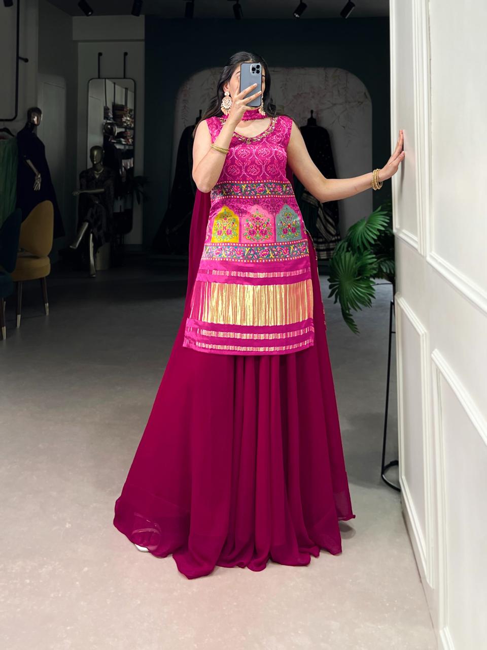 Model showcasing a premium pink Bandhani print sleeveless tunic kurti with multi-color traditional arch patchwork embroidery, heavy gold fringe hem, and solid flared pink georgette sharara pants inside a boutique. Main image